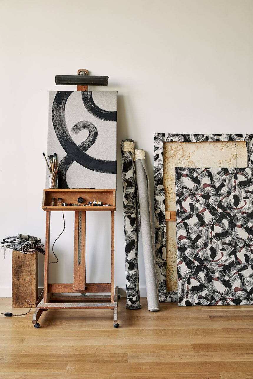 An easel with a painting showing swirling black lines sits beside fabric covered canvases.
